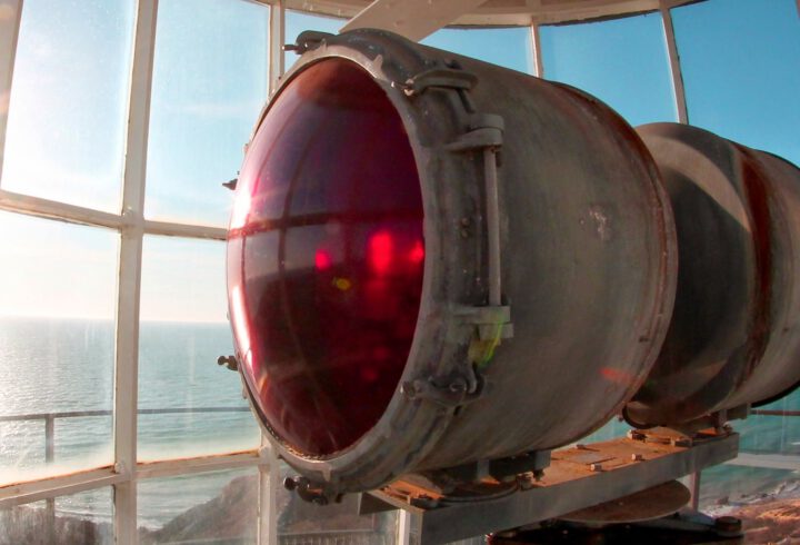 Large red lighthouse lens inside a lantern room overlooking the ocean.