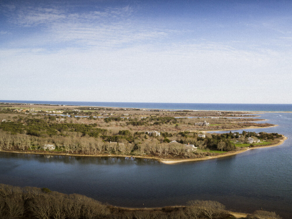 Aerial view of a coastal landscape of Martha's Vineyard with calm blue water, wooded shoreline, scattered homes, and the ocean in the distance.