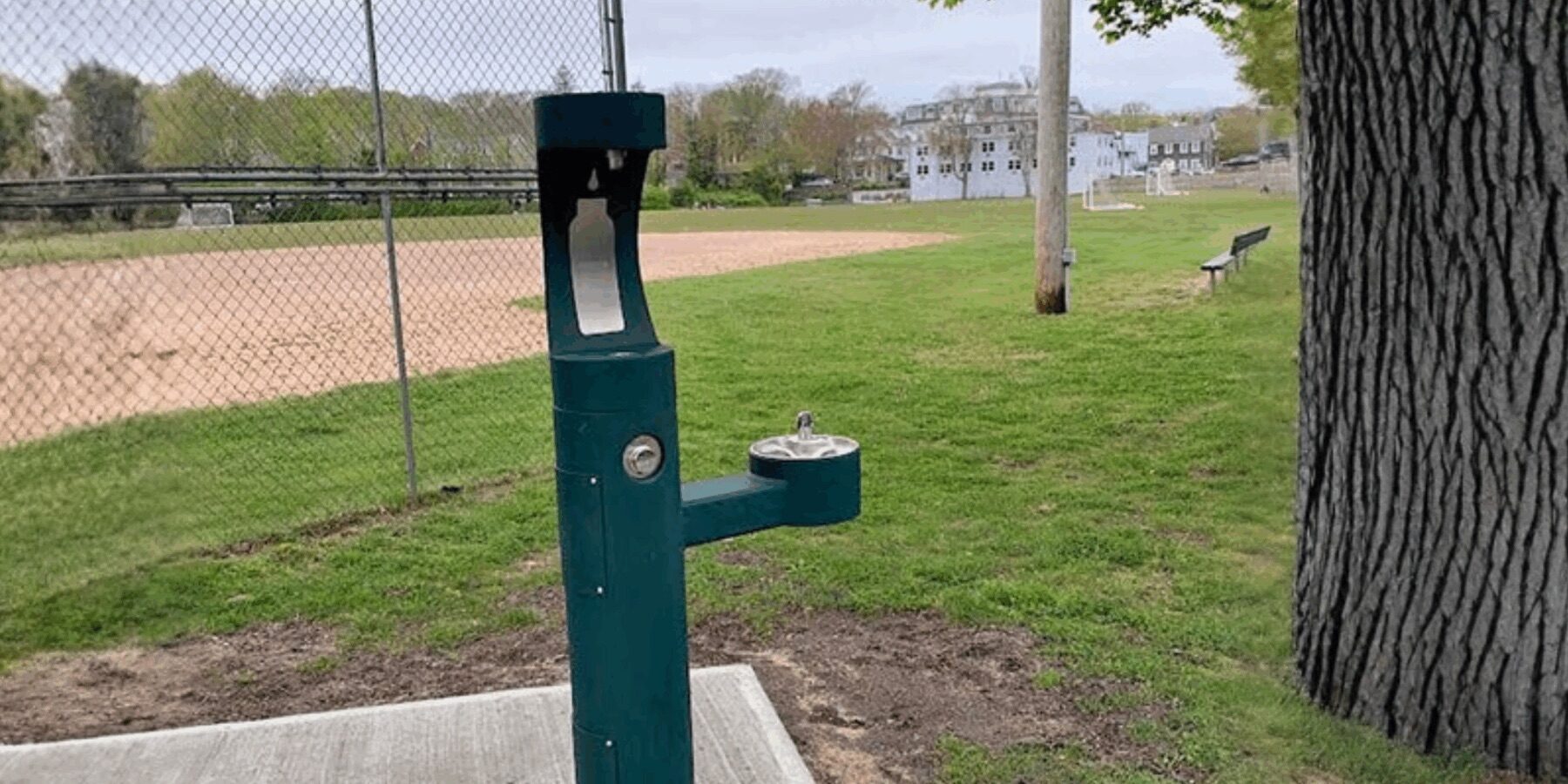 A public water bottle filling station and drinking fountain stand beside a fenced baseball field and grassy park.
