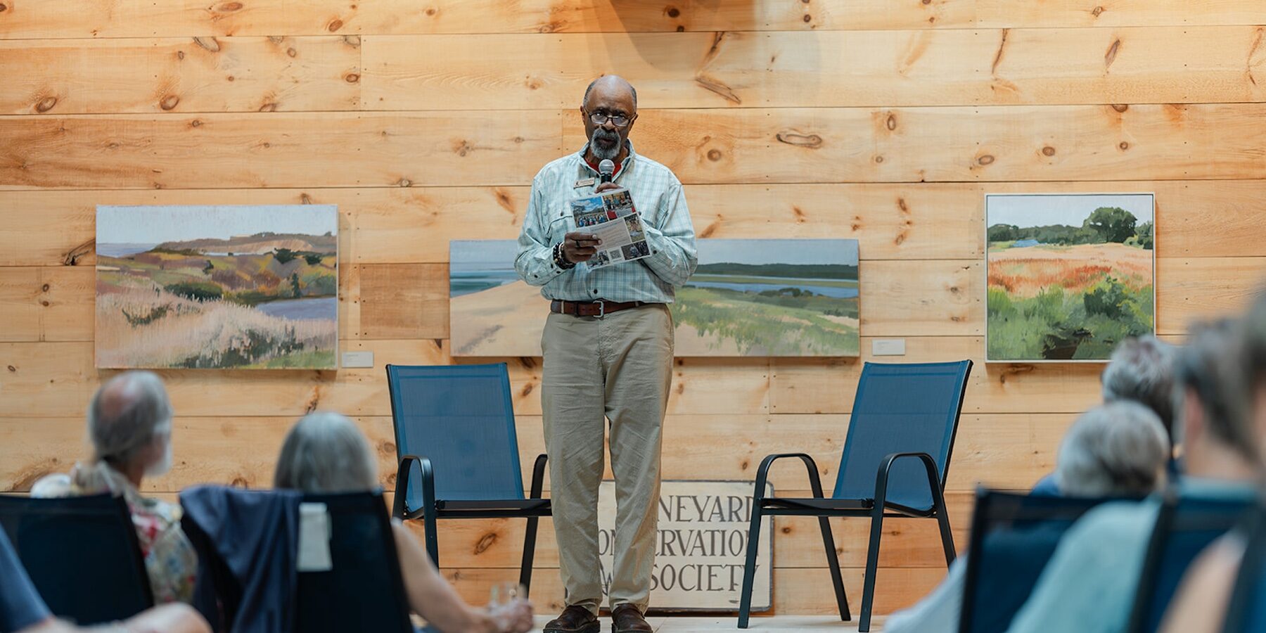 Speaker addressing an audience at a Vineyard Conservation Society event with landscape paintings displayed behind him.