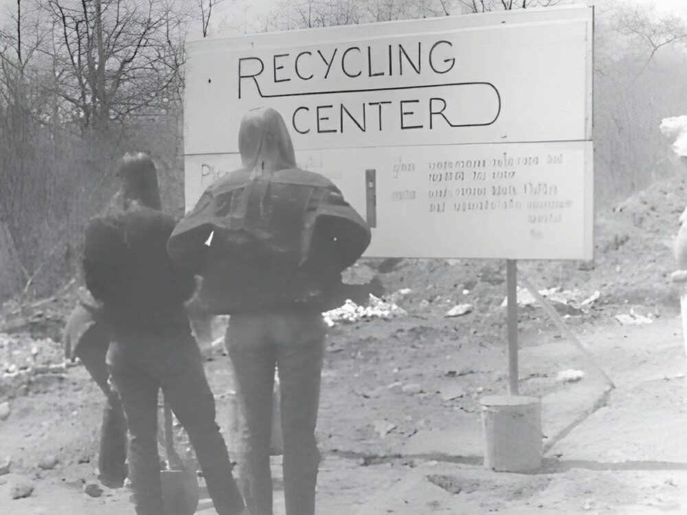 Black-and-white photo of two people standing beside a large sign reading “Recycling Center” in an outdoor, wooded area.