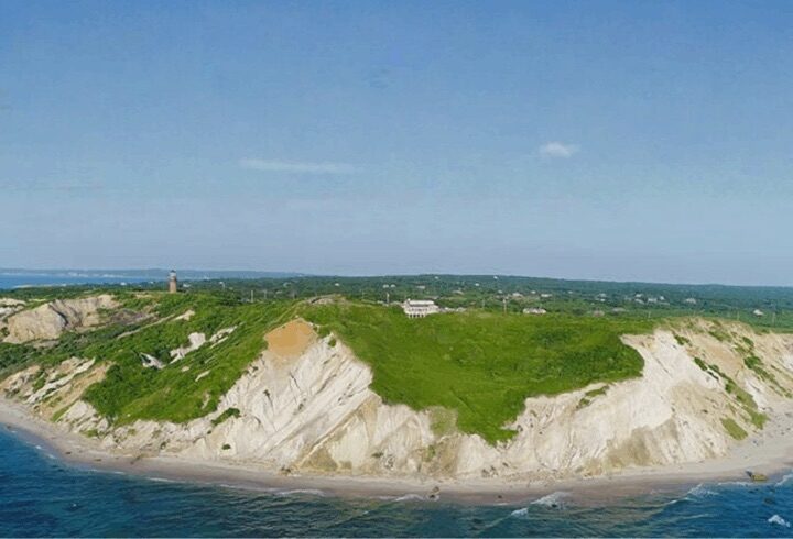 A wide coastal view shows steep white cliffs topped with green grass and scattered buildings beside the blue ocean.