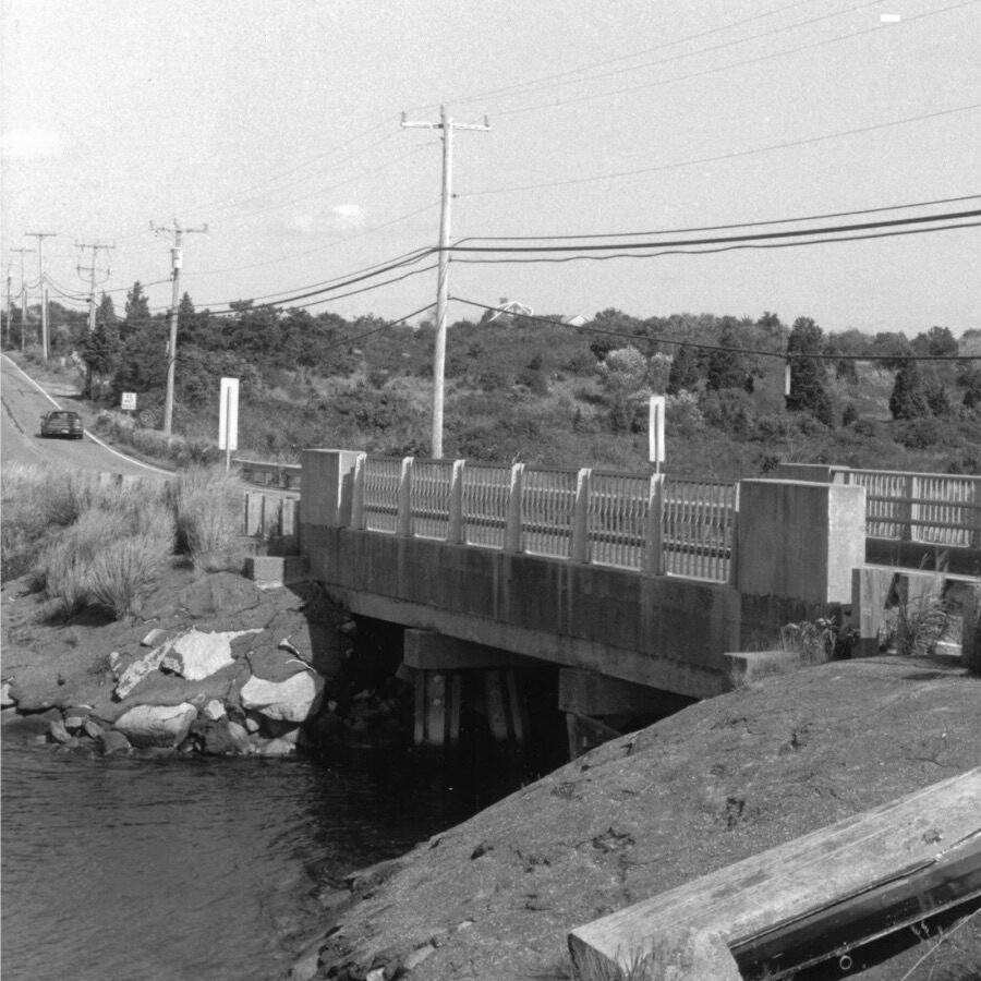Black-and-white photo of an old bridge in Oak Bluffs crossing a narrow waterway beside a road lined with utility poles and wires.