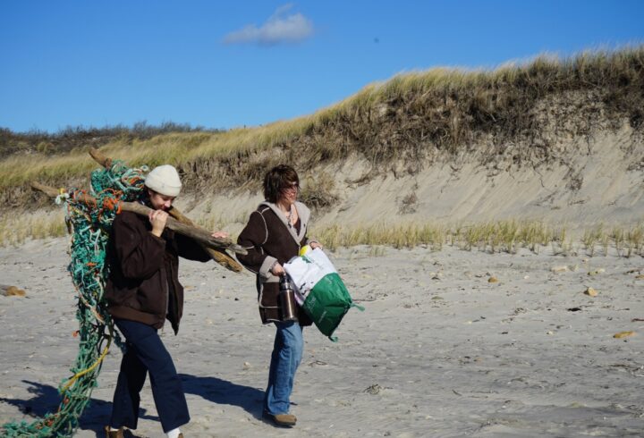 wo people walk along a sandy beach near grassy dunes, carrying a large piece of driftwood tangled with rope and a bag of collected debris.