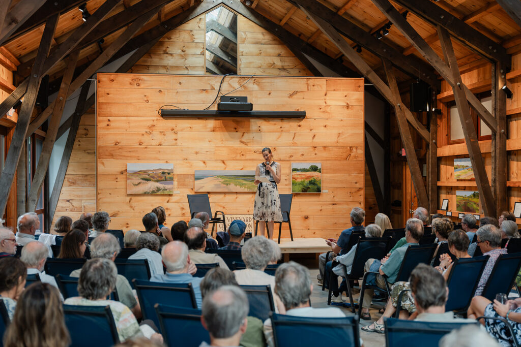 A speaker stands on a stage inside a wooden hall, addressing a seated audience during a community talk or event.
