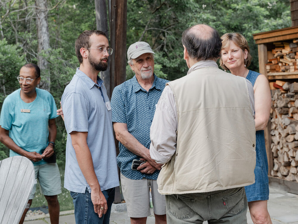 A small group of adults stands outdoors talking together on a patio beside stacked firewood and trees.